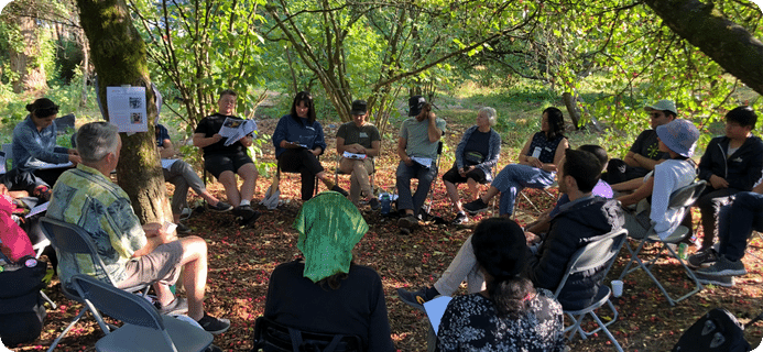 Attendees of the annual general meeting are surrounded by trees and seated on chairs in a circle.