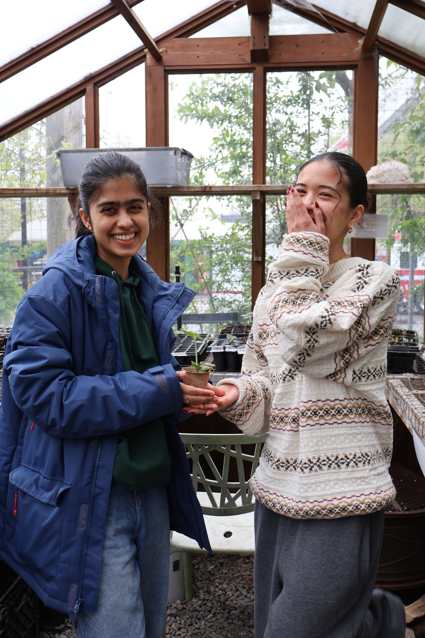 Youth participants Vaibhavi and Arianne in the greenhouse at Strathcona Community Gardens, holding up a potted plant and smiling.