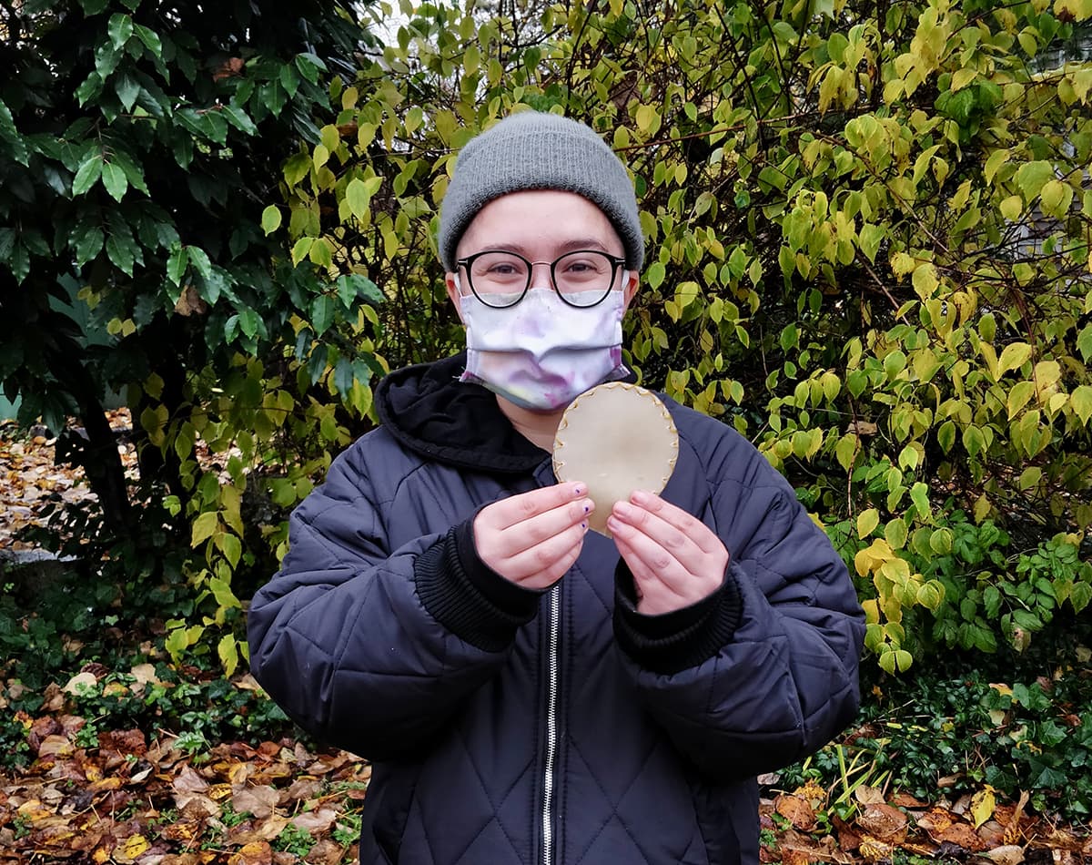 Portrait of a youth wearing a grey beanie, glasses and a mask holding a beige circular piece of woven leather.