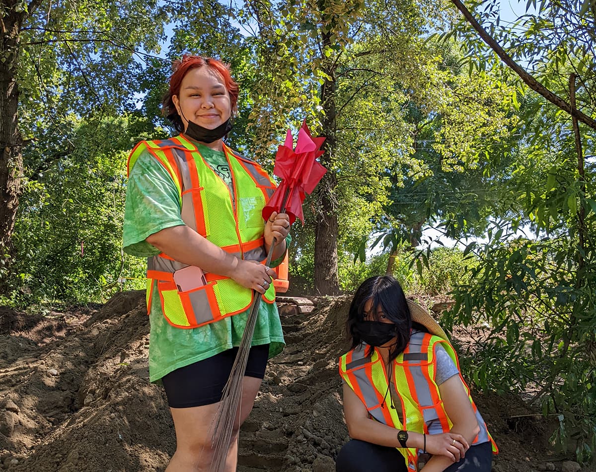 Two young women wearing high visibility vests are standing and kneeling outside near trees. One of them is holding metal pegs with red flags at the top of them.