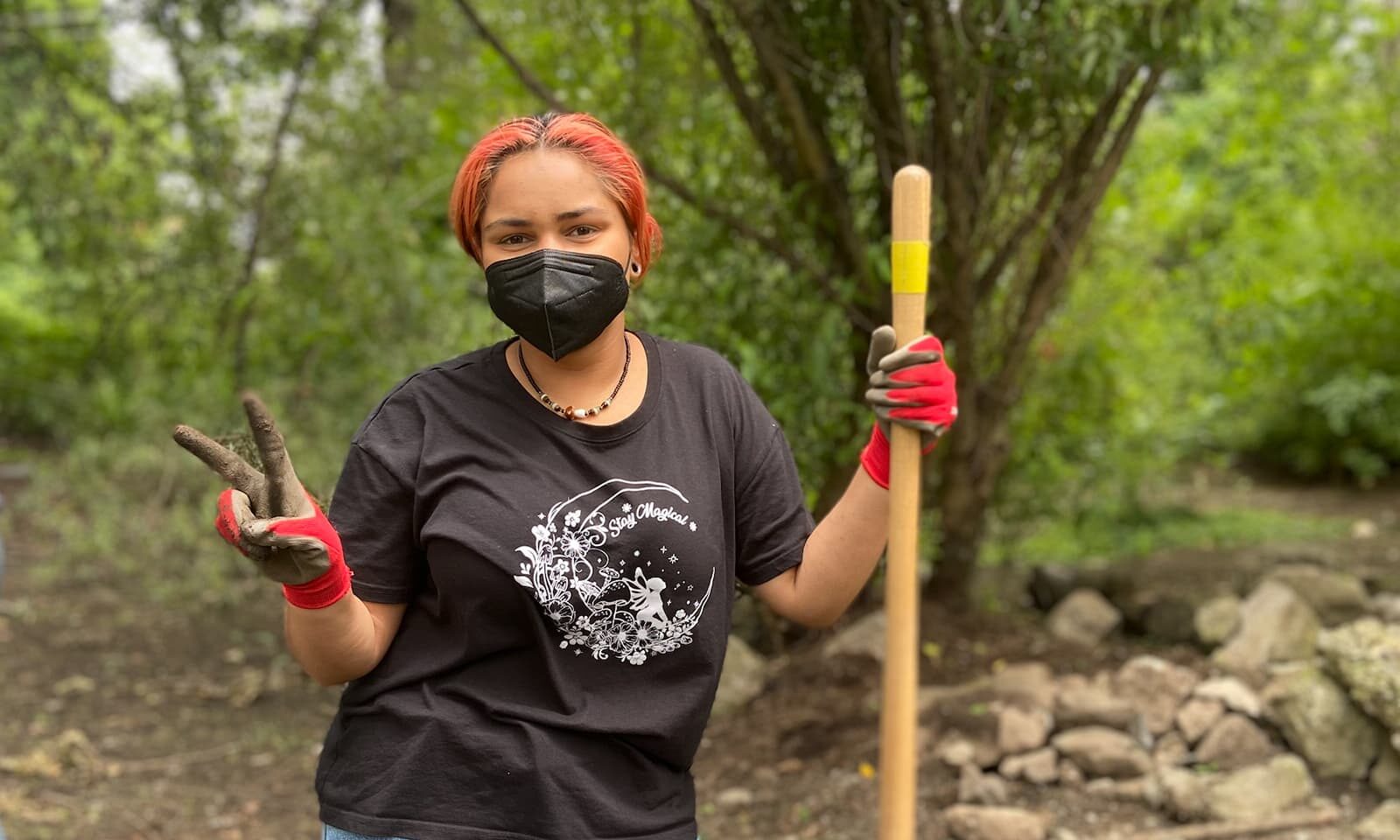 A young woman is standing outside holding a wooden tool for tending to a garden. She is wearing a mask and gardening gloves and is putting up a piece sign with one hand.