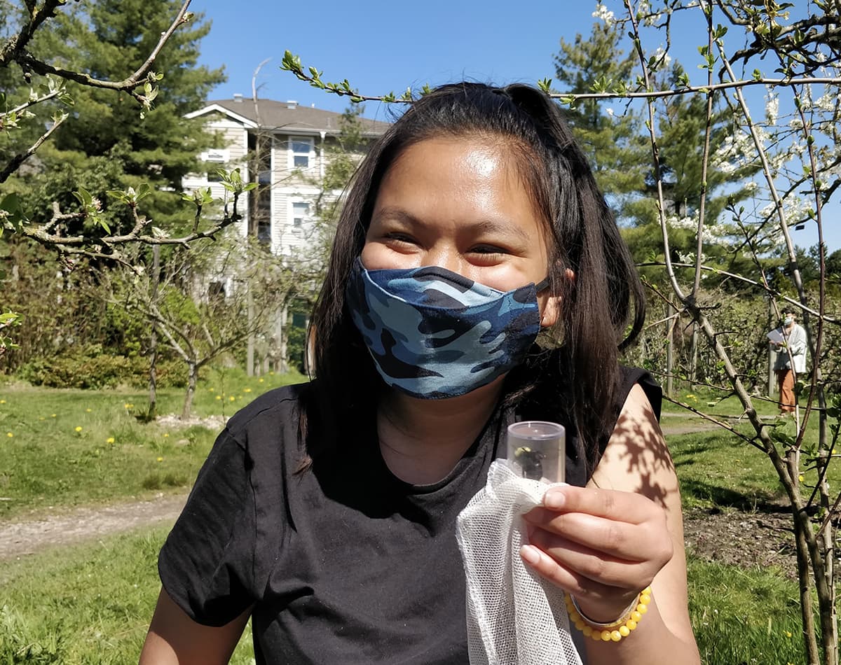 A young woman is outside and holding a small plastic container which has a bumblebee inside.