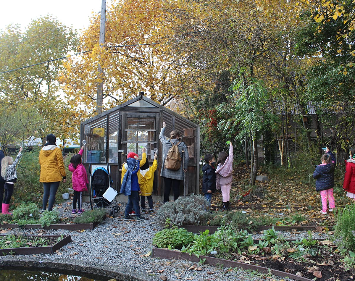 A group of children and two adults are standing around a garden and greenhouse with their hands raised.