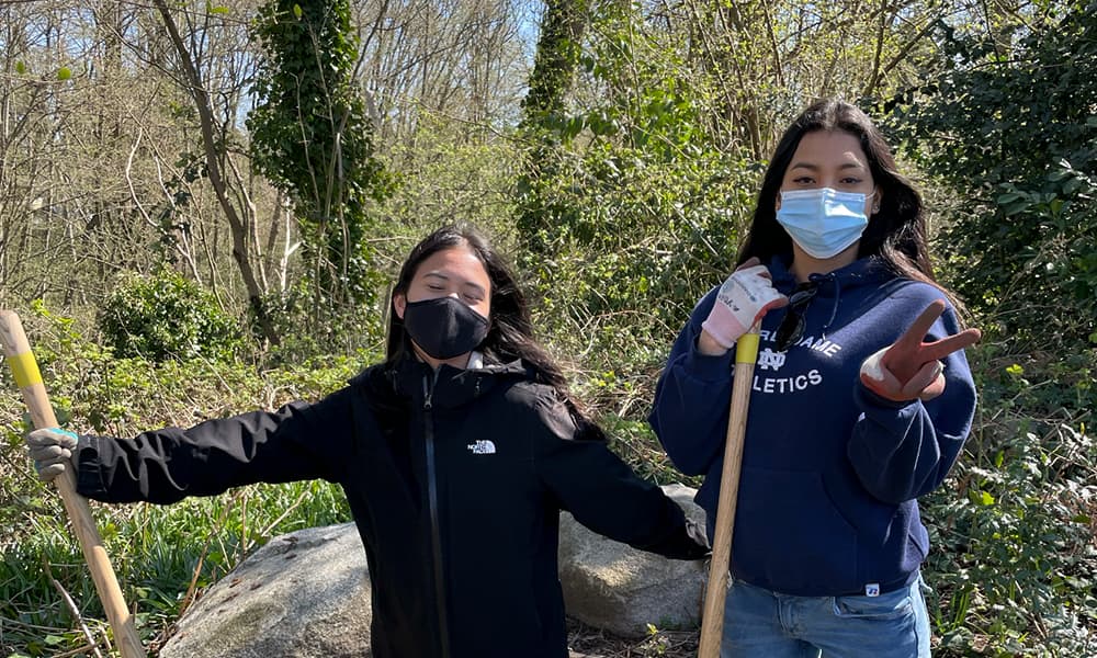 Two young women are wearing masks and standing outside. They are wearing gardening gloves and holding a gardening tool.