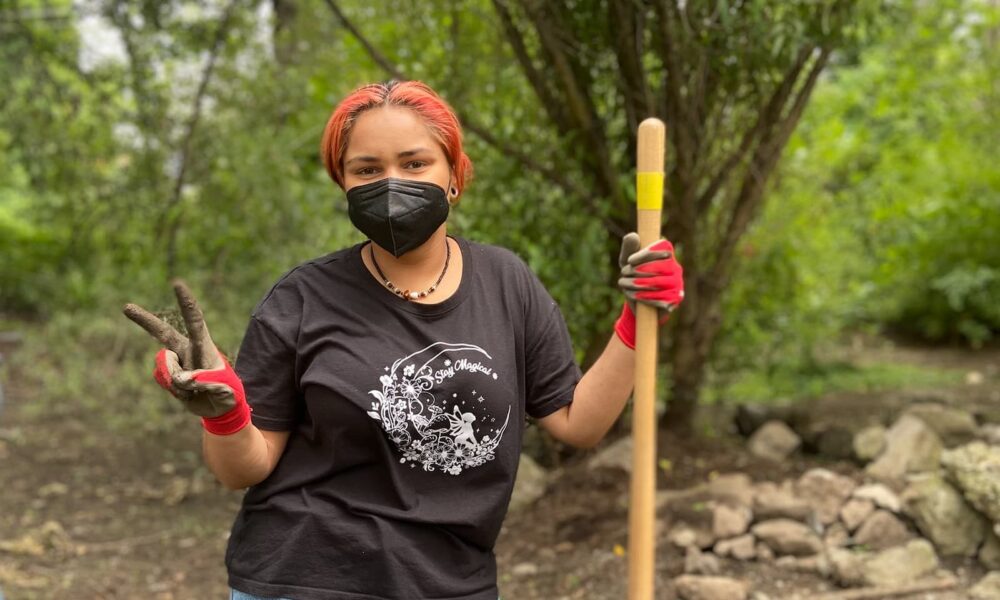 A young person is standing outside holding a wooden tool for tending to a garden. They are wearing a mask and gardening gloves and are putting up a piece sign with one hand.