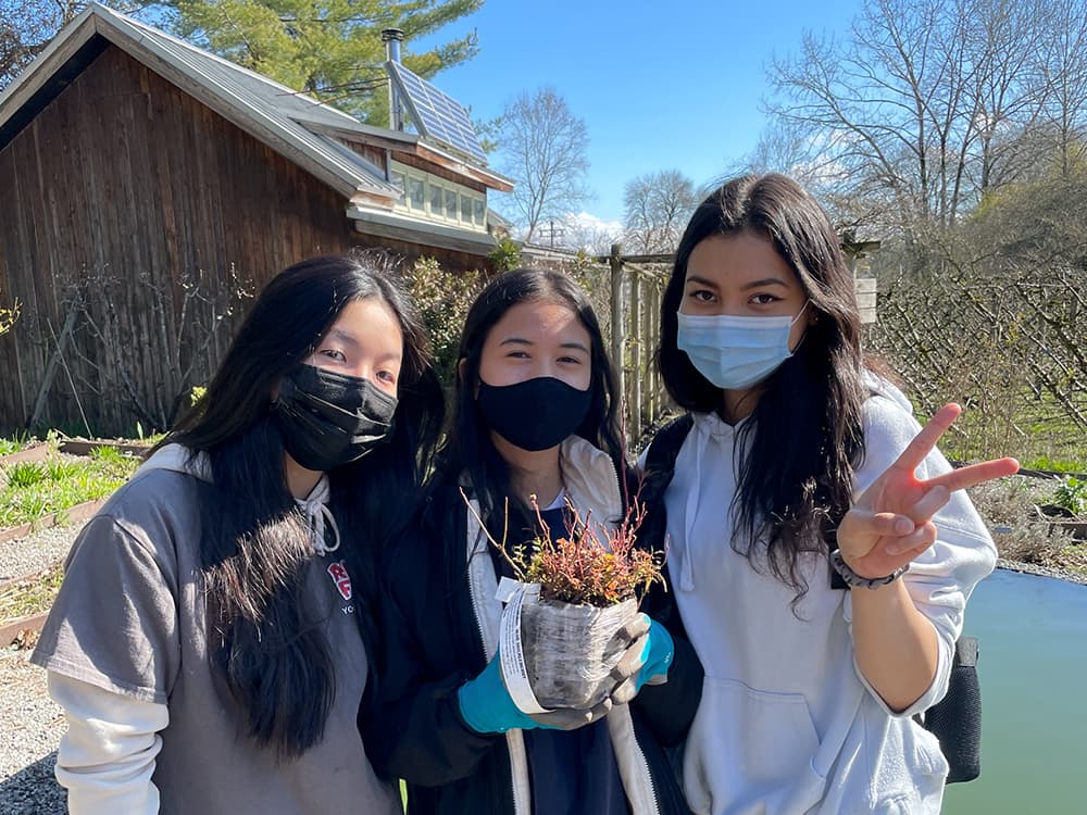 Three young women are standing outside at the Strathcona Community Garden posing while holding a plant in a nursery pot.