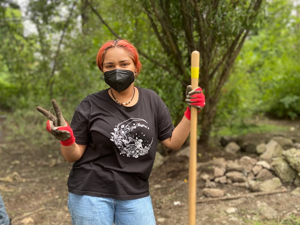 A young woman is standing outside holding a wooden tool for tending to a garden. She is wearing a mask and gardening gloves and is putting up a piece sign with one hand.