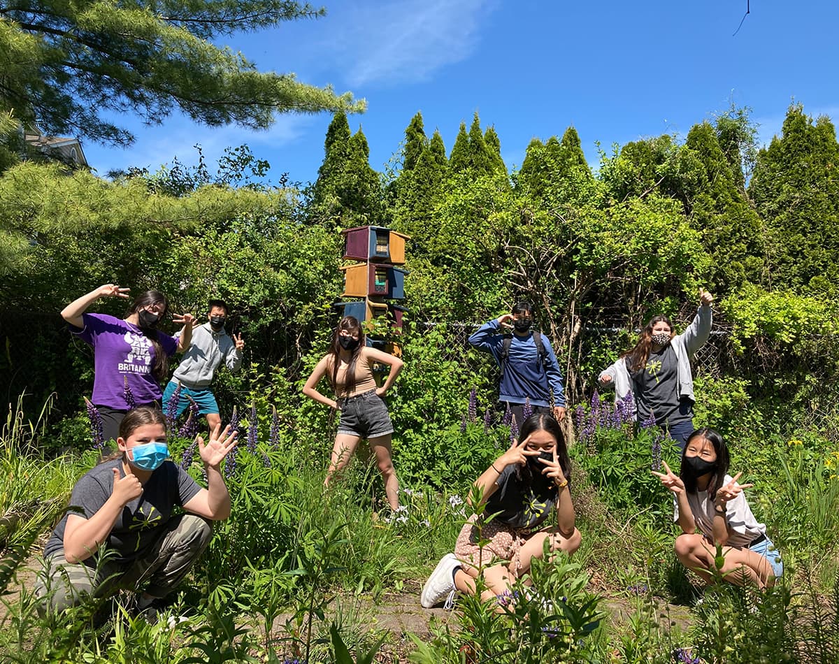 A group of youth are standing and kneeling outside in a green field surrounded by trees. They are making gestures with their hands and are happily posing.