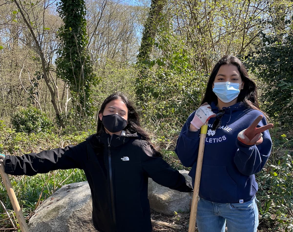 Two young women are wearing masks and standing outside. They are wearing gardening gloves and holding a gardening tool.