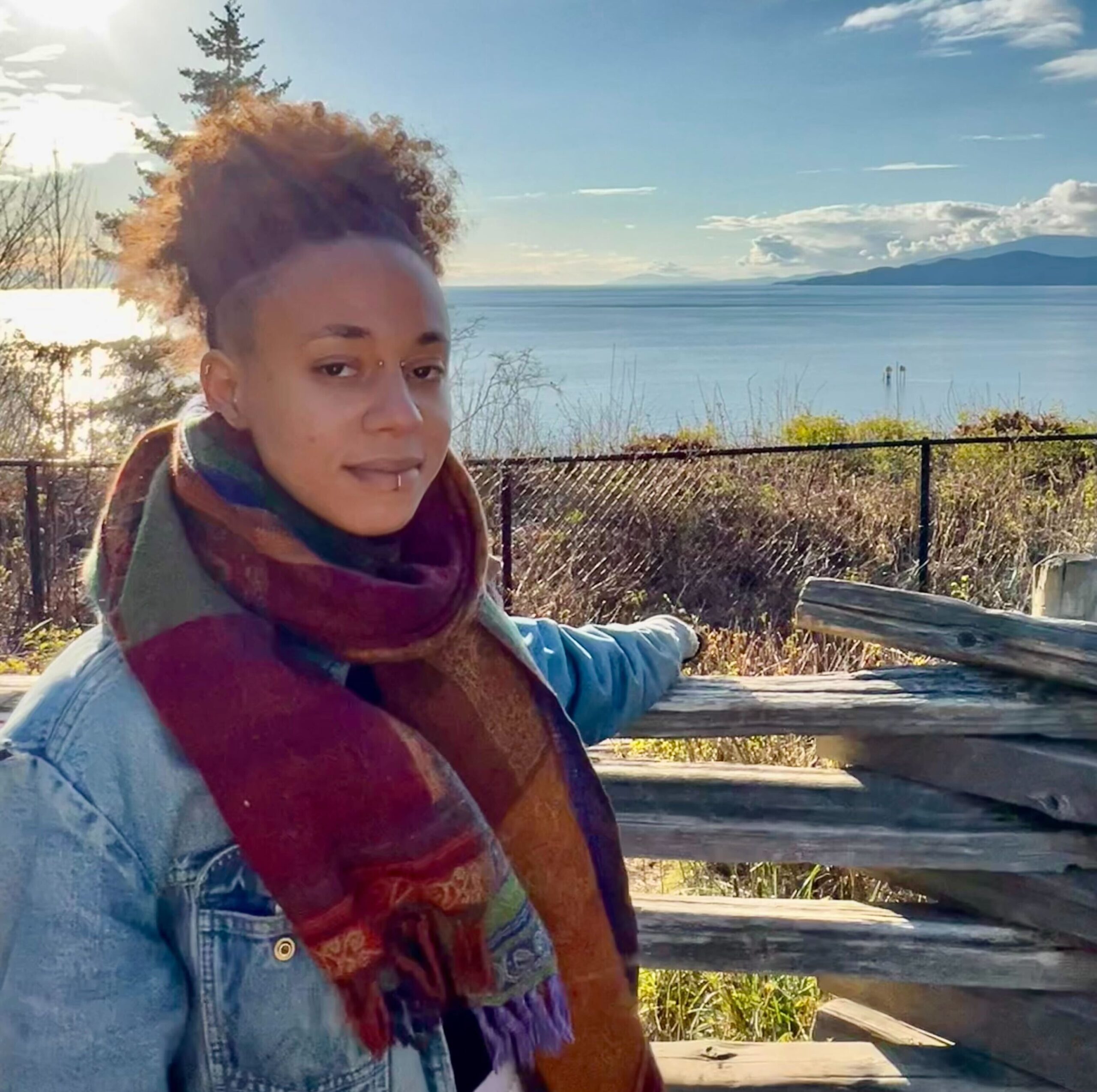 Savannah is wearing a denim jacket and large scarf. They have one arm on a wooden fence with grasses and the ocean in the background.