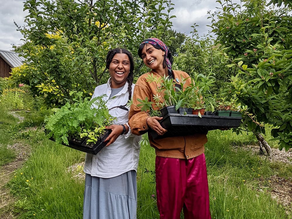 Two smiling women are standing outside surrounded by trees. They are holding bins that contain plants in nursery pots.