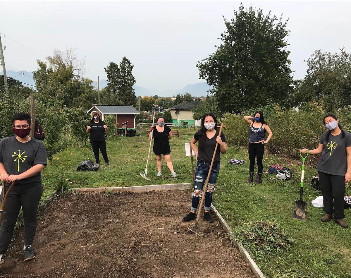 A group of youth are standing outside holding various gardening tools like rakes and shovels. Some of them are wearing t-shirts with the EYA dandelion icon.