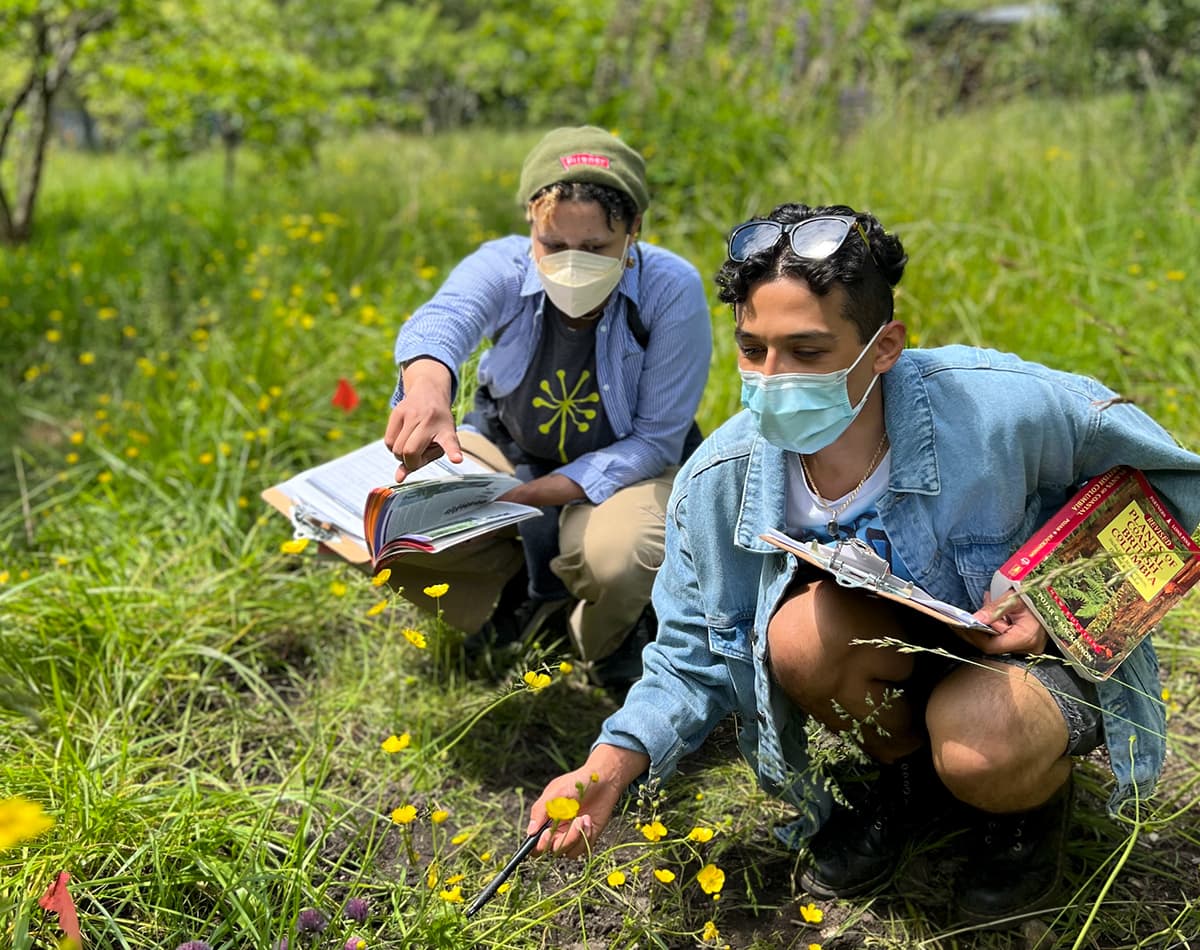 Two youth are kneeling outdoors while holding plant identification books and clip boards and pointing at different plant life.