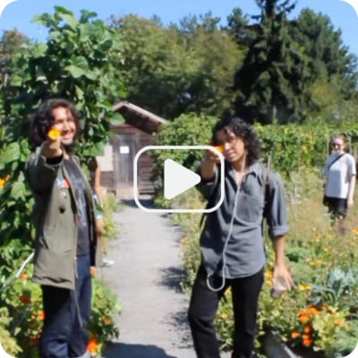 Two youth face the camera holding a yellow calendula flower. A video play button is in the centre of the image