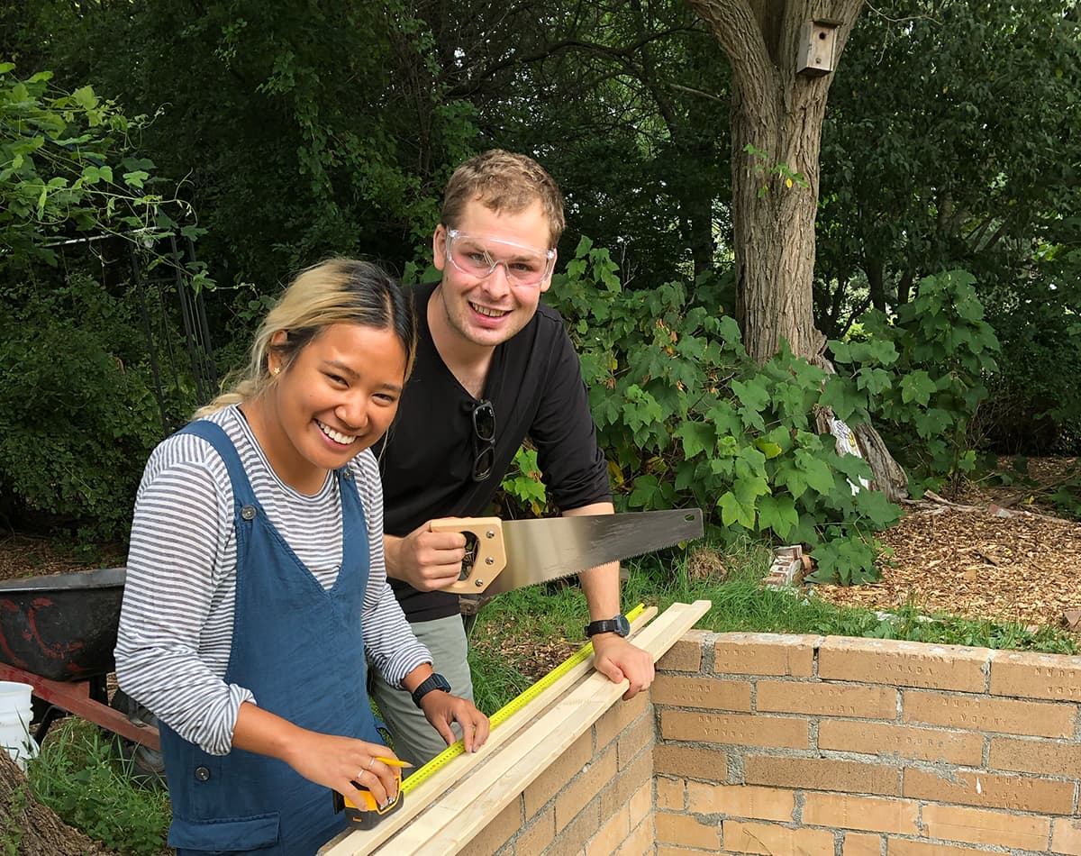 A young woman and man are outside and using tools to build a brick structure.